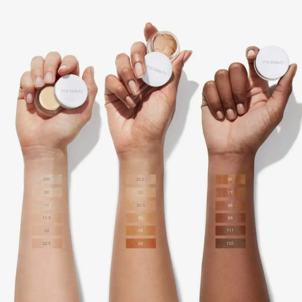 A hand applying RMS Beauty concealer with the product shown on a clean, white background
